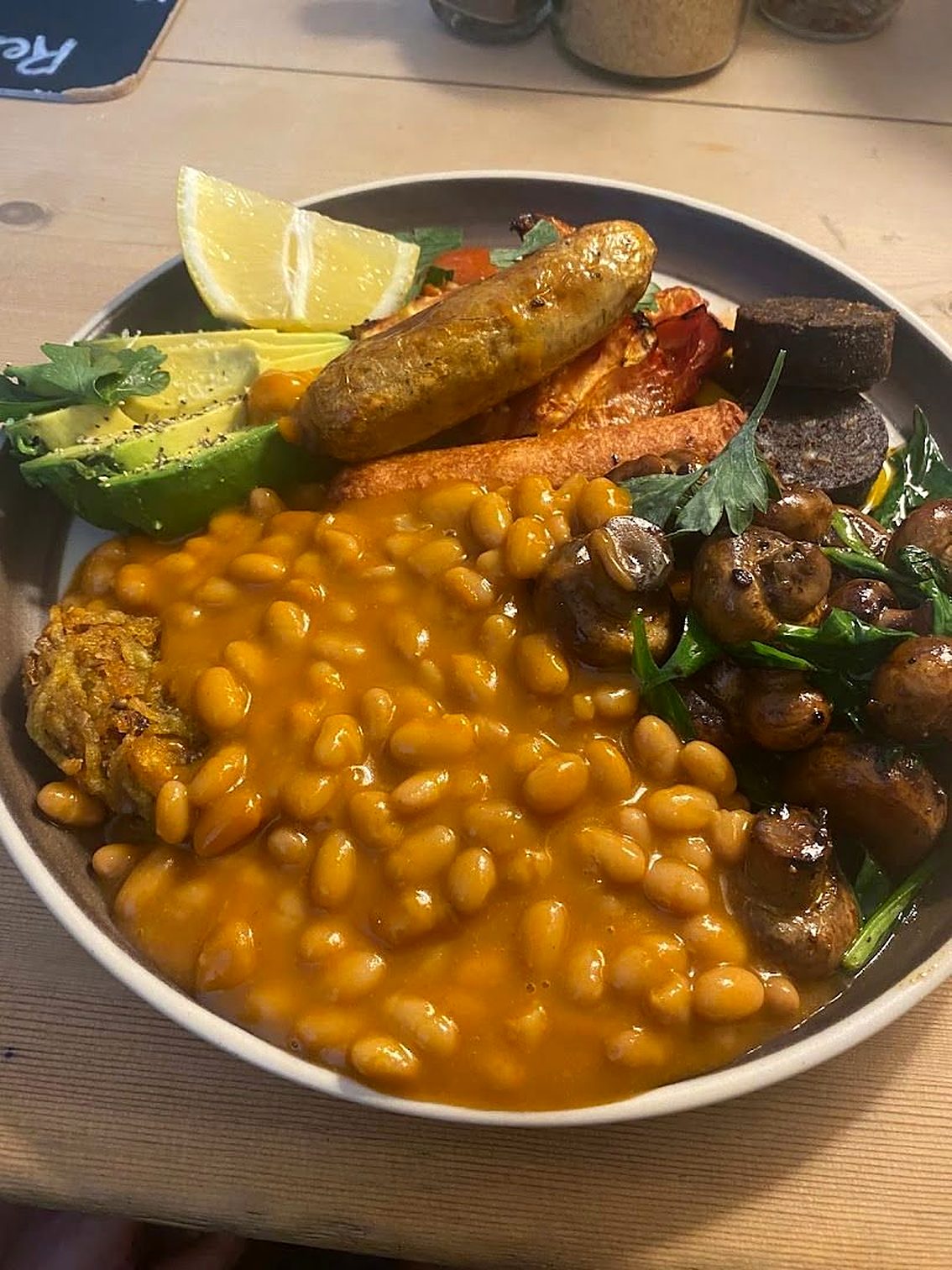 A breakfast bowl: beans, a sausage, avocado, mushrooms, roasted vegetables and a wedge of lemon