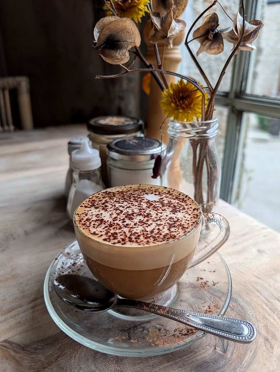 A cappuccino dusted with cocoa in a glass cup on a wooden table, dried flowers in a jar beside it