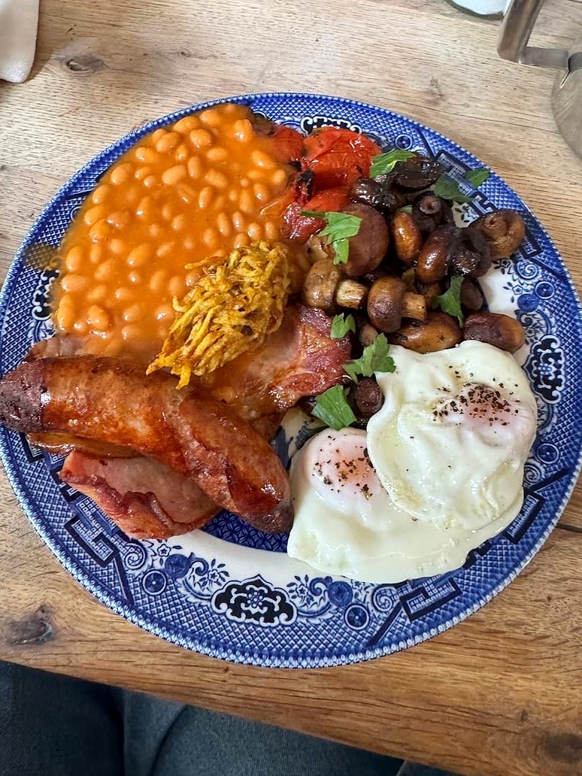 A full English breakfast on a blue-and-white patterned plate: sausage, bacon, two fried eggs, beans, mushrooms and tomatoes