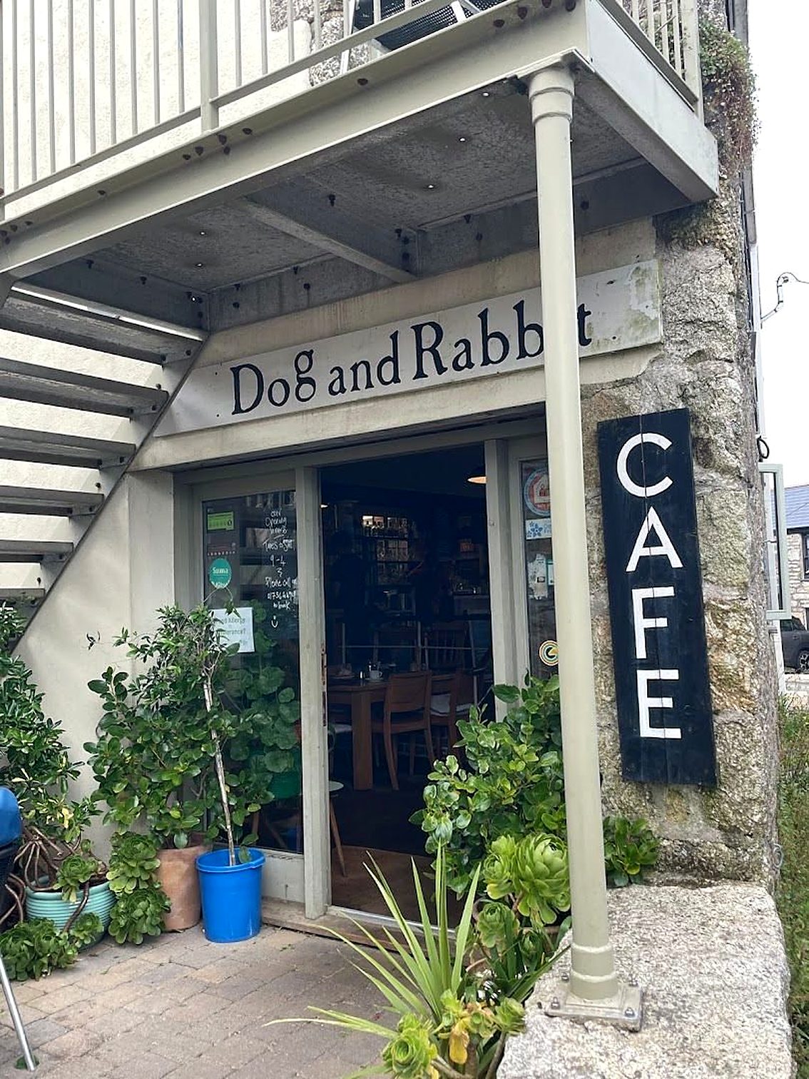 The café on North Row, St Just: a “Dog and Rabbit” sign over the door, a tall black “CAFE” board on the granite wall, and pots of plants by the entrance